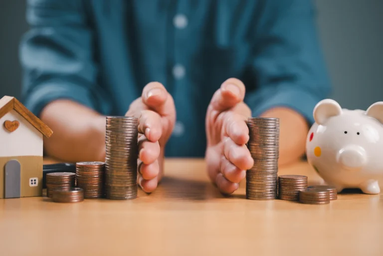 A persons hands part piles of coins surrounded by a model house and piggy bank on a table